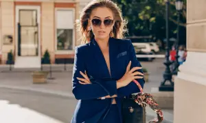 Elegancka kobieta w granatowym garniturze i okularach przeciwsłonecznych, z torebką przewieszoną przez ramię. Tak może wyglądać dziennikarka w drodze na ważne spotkanie.