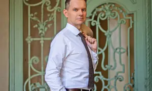 Tomasz Roy Szabelewski w białej koszuli i krawacie, z marynarką na ramieniu, przed ozdobnymi drzwiami.