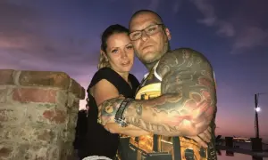 Para, przytulająca się na tle zachodzącego słońca. Ona, jak wygląda żona Popka, ma blond włosy i czarną bluzkę. On, wytatuowany mężczyzna w okularach, obejmuje ją.