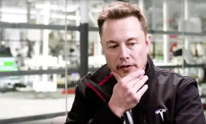 Elon Musk w czarnej kurtce z logo Tesli, z ręką na brodzie, w tle fabryka. Jego wizja przyszłości, niczym z tau techno mechanicus, inspiruje.
