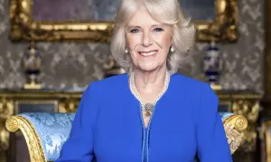 Uśmiechnięta Camilla Parker Bowles w niebieskiej sukience i perłach, siedząca na ozdobnym fotelu.