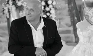 Jacek, znany jako królowe życia, w eleganckim garniturze, z uśmiechem spogląda w obiektyw. W tle widać suknię ślubną.
