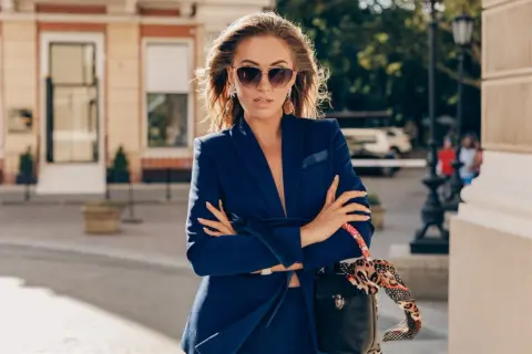 Elegancka kobieta w granatowym garniturze i okularach przeciwsłonecznych, z torebką przewieszoną przez ramię. Tak może wyglądać dziennikarka w drodze na ważne spotkanie.