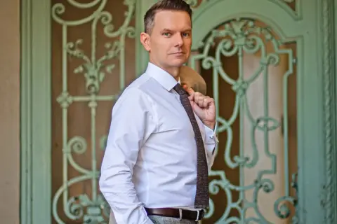 Tomasz Roy Szabelewski w białej koszuli i krawacie, z marynarką na ramieniu, przed ozdobnymi drzwiami.
