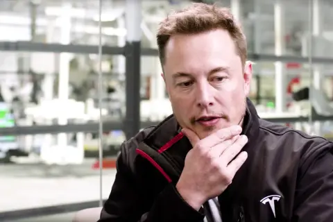 Elon Musk w czarnej kurtce z logo Tesli, z ręką na brodzie, w tle fabryka. Jego wizja przyszłości, niczym z tau techno mechanicus, inspiruje.