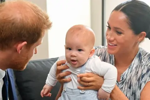 Meghan Markle trzyma swoje dziecko, które z zaciekawieniem patrzy na księcia Harry'ego.