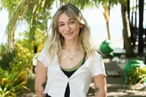 Uśmiechnięta Fabienne Marta Wiśniewska w białej bluzce na tle tropikalnej roślinności i plaży.