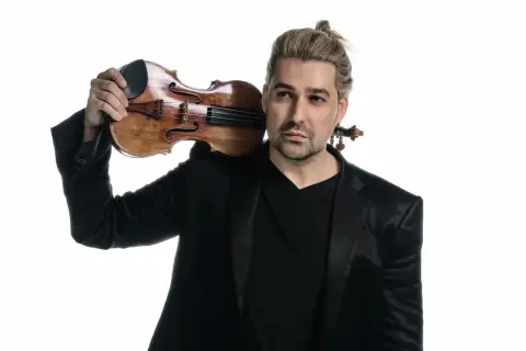 David Garrett z blond włosami spiętymi w kucyk, w czarnym garniturze, trzymający skrzypce na ramieniu.