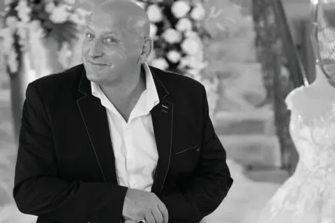 Jacek, znany jako królowe życia, w eleganckim garniturze, z uśmiechem spogląda w obiektyw. W tle widać suknię ślubną.