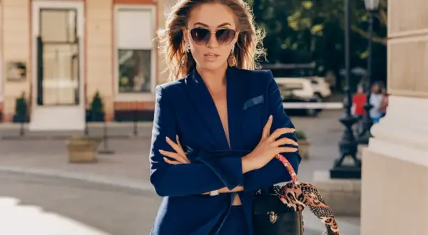 Elegancka kobieta w granatowym garniturze i okularach przeciwsłonecznych, z torebką przewieszoną przez ramię. Tak może wyglądać dziennikarka w drodze na ważne spotkanie.