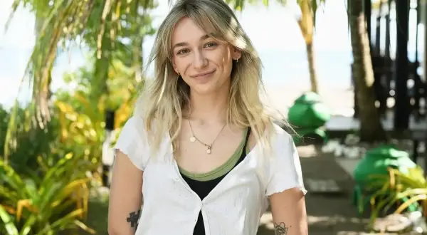 Uśmiechnięta Fabienne Marta Wiśniewska w białej bluzce na tle tropikalnej roślinności i plaży.