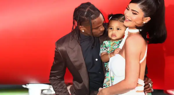 Kylie Jenner z dzieckiem i Travisem Scottem na tle czerwonej ściany.