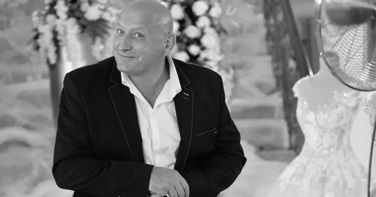 Jacek, znany jako królowe życia, w eleganckim garniturze, z uśmiechem spogląda w obiektyw. W tle widać suknię ślubną.