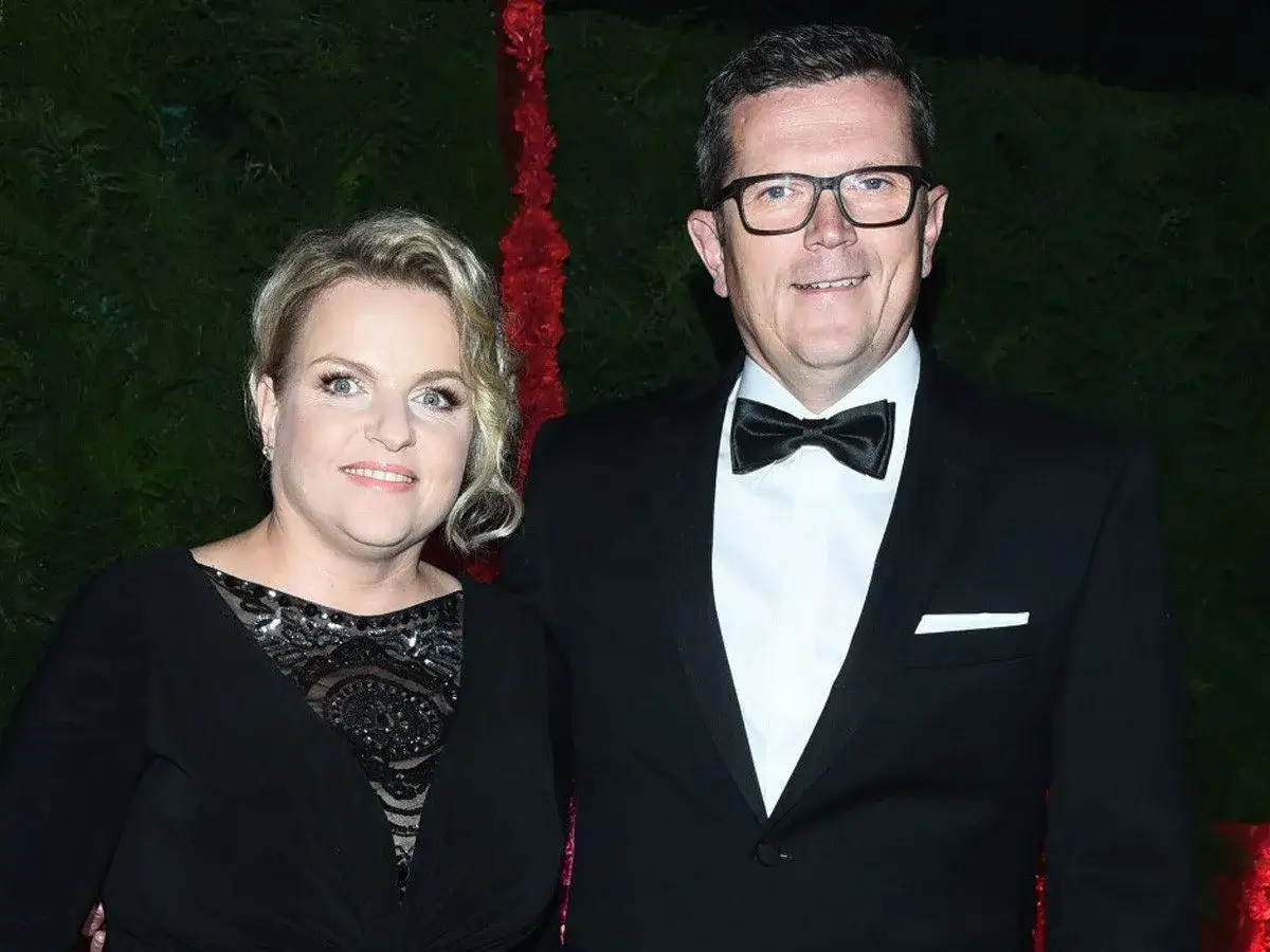 Marcin Bosacki z nową partnerką, uśmiechnięci i elegancko ubrani, pozują do zdjęcia na tle zieleni i czerwonych dekoracji.