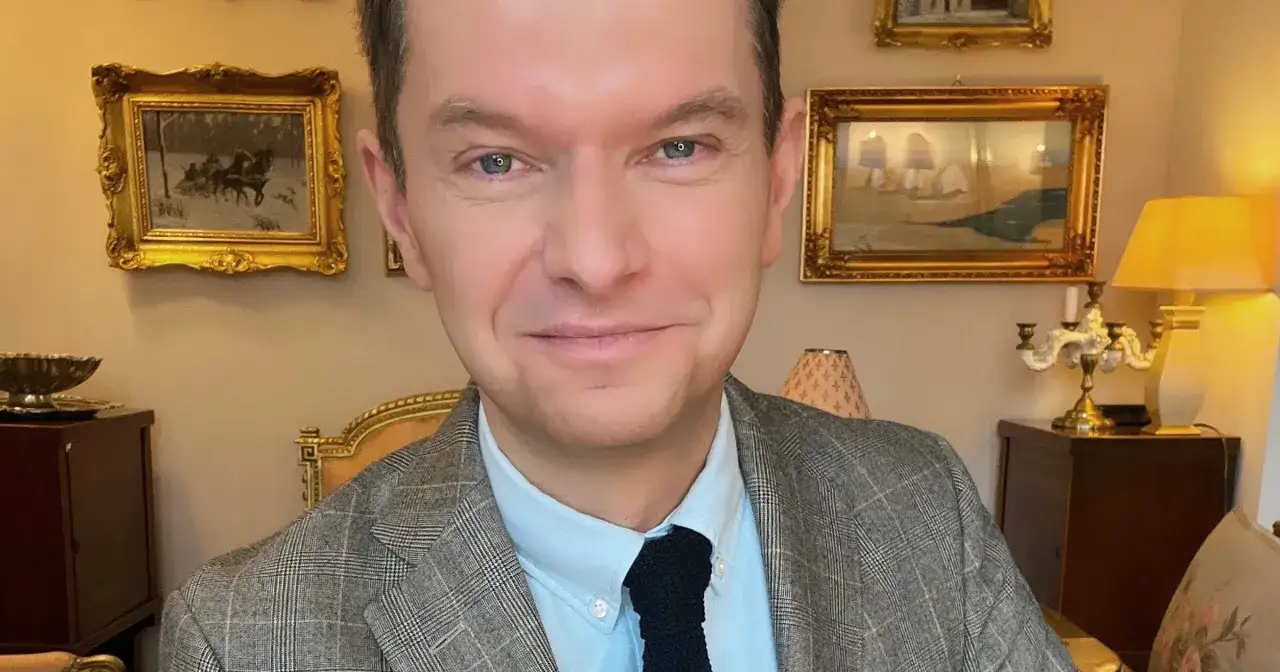 Tomasz Roy Szabelewski w eleganckim garniturze, na tle obrazów i antyków.