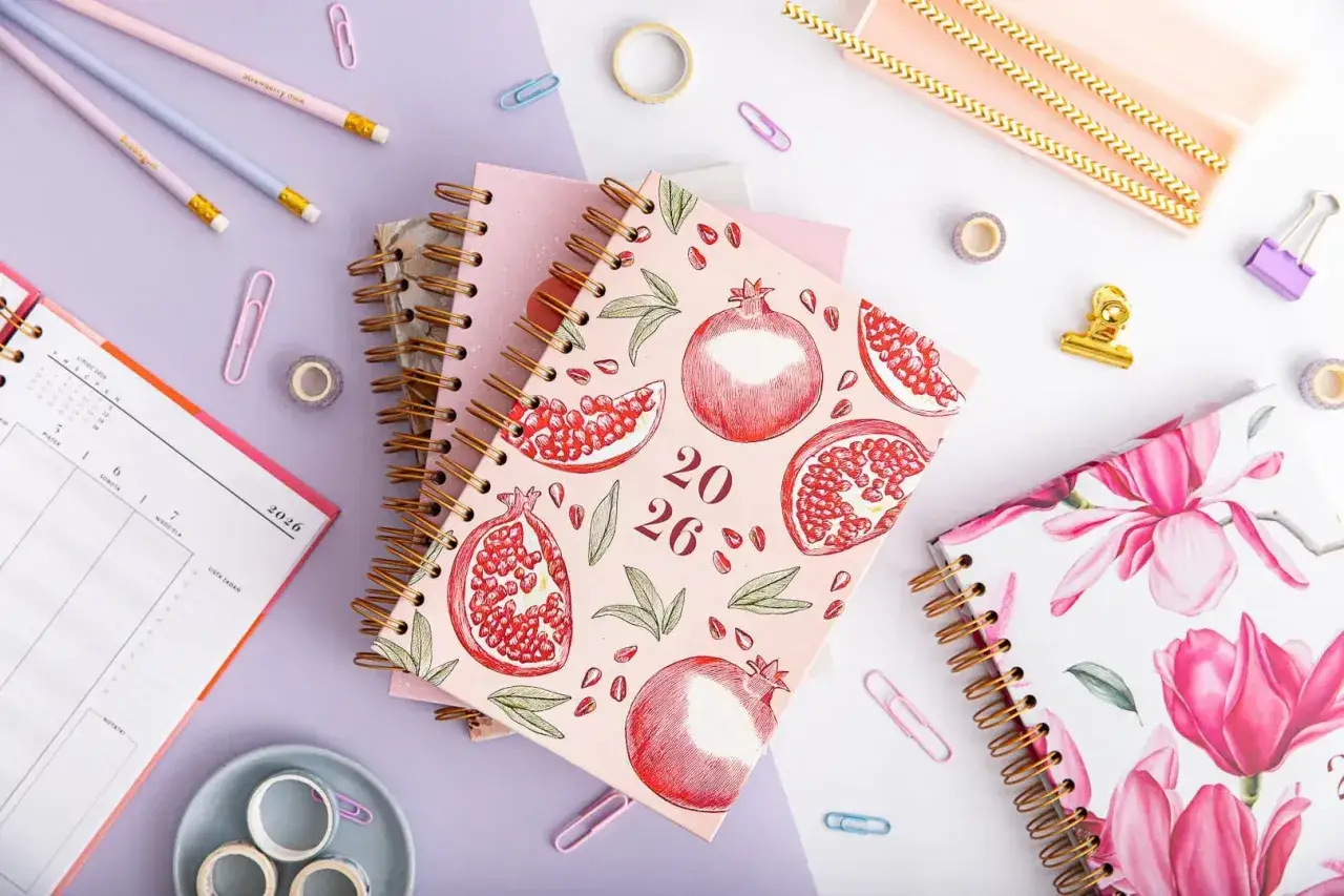 Czy planner i kalendarz od influencerki pomagają w organizacji czasu? Na zdjęciu widać stylowe notesy z motywem granatów i magnolii, obok ołówków i spinaczy.