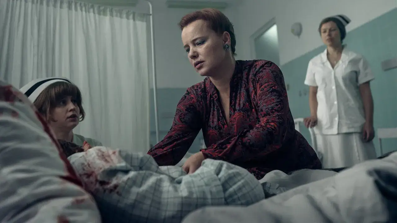 Katarzyna Warnke, która polska aktorka dostała rolę w wielkiej produkcji Netflixa, pochyla się nad pacjentem w szpitalu. Obok stoi pielęgniarka.