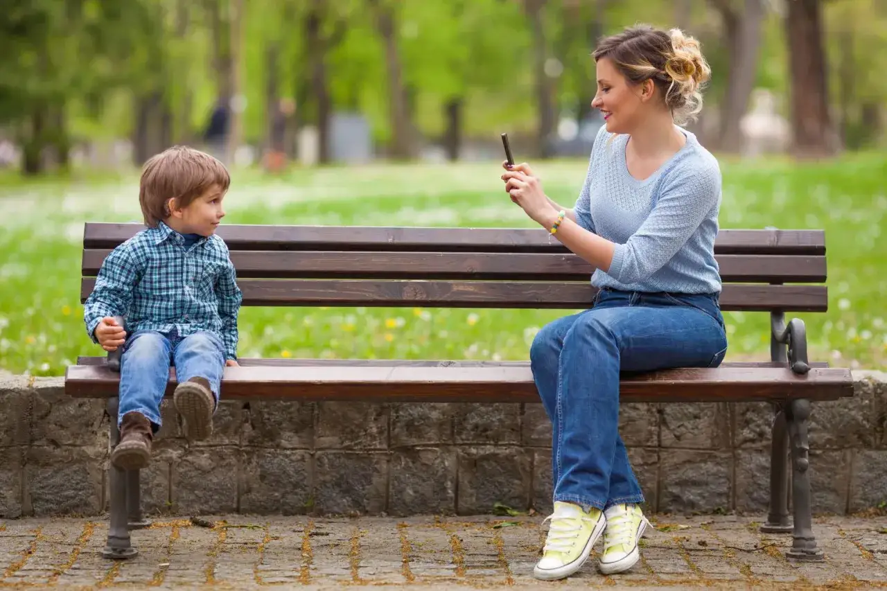 Kobieta robi zdjęcie dziecku na ławce w parku. Zastanawiamy się, dlaczego gwiazdy decydują się na ukrywanie wizerunku dzieci w sieci.