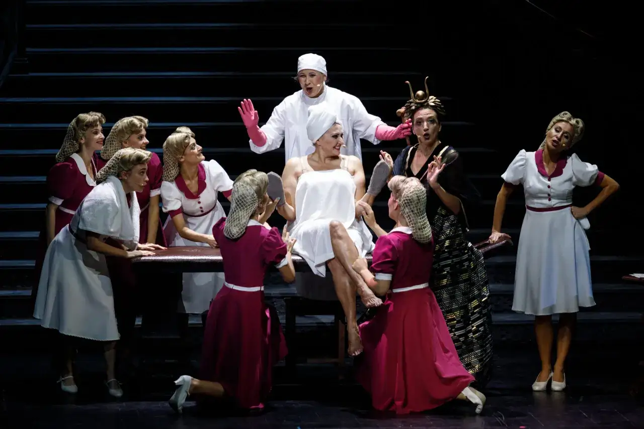 Scena z opery. Czy warto kupić bilet na spektakl teatralny z obsadą samych celebrytów? Grupa kobiet w strojach z epoki, jedna w ręczniku na głowie, druga w czepku.