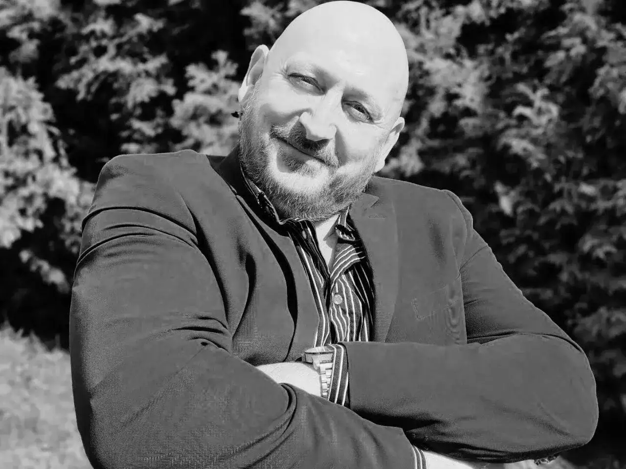 Jacek, król życia, w garniturze i pasiastym krawacie, z rękami skrzyżowanymi na piersi, uśmiecha się do aparatu na tle drzew.