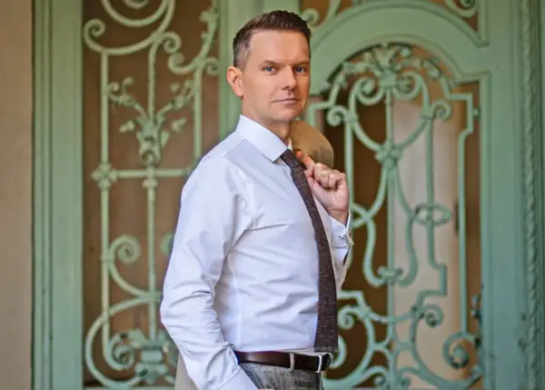 Tomasz Roy Szabelewski w białej koszuli i krawacie, z marynarką na ramieniu, przed ozdobnymi drzwiami.