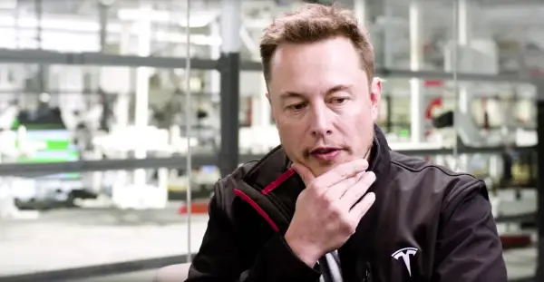 Elon Musk w czarnej kurtce z logo Tesli, z ręką na brodzie, w tle fabryka. Jego wizja przyszłości, niczym z tau techno mechanicus, inspiruje.