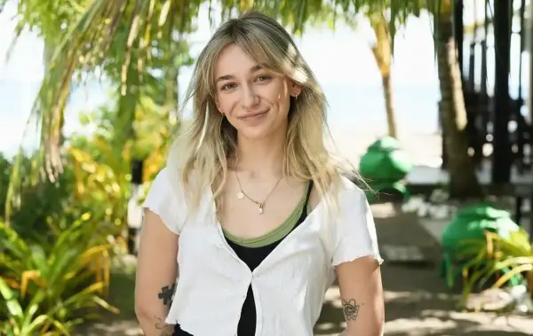 Uśmiechnięta Fabienne Marta Wiśniewska w białej bluzce na tle tropikalnej roślinności i plaży.