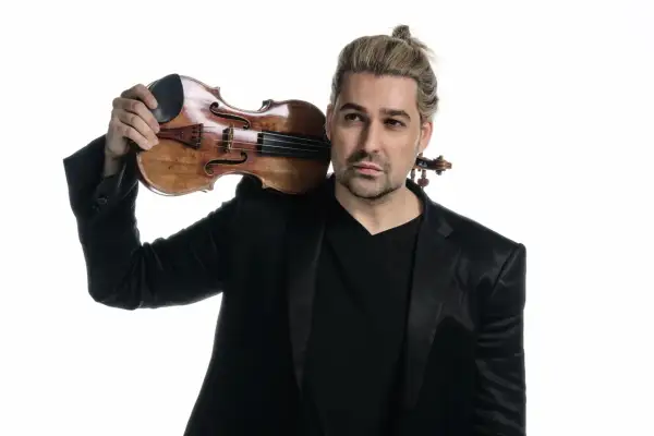 David Garrett z blond włosami spiętymi w kucyk, w czarnym garniturze, trzymający skrzypce na ramieniu.