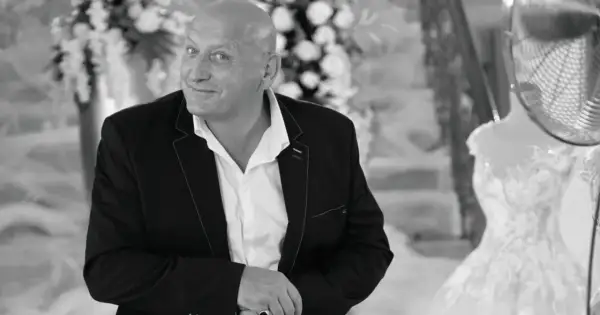 Jacek, znany jako królowe życia, w eleganckim garniturze, z uśmiechem spogląda w obiektyw. W tle widać suknię ślubną.